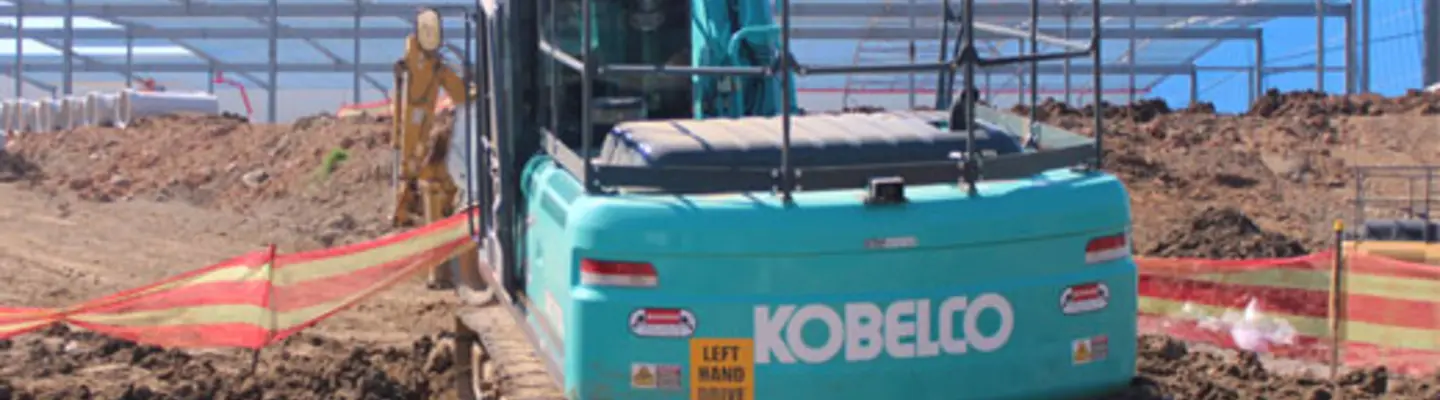 Excavator digging at an active construction site, with safety barriers and warning signs visible around the work area.