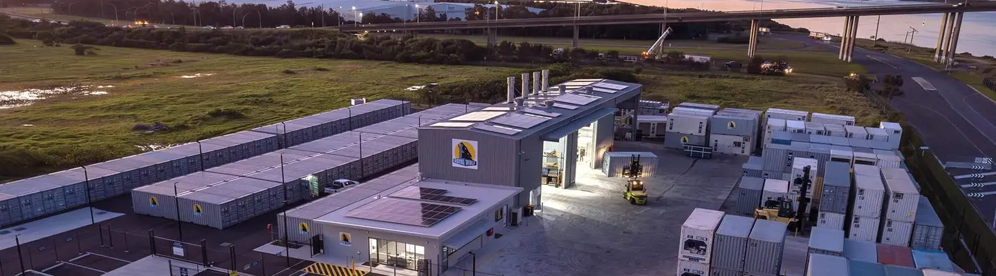 Aerial view of a container facility and warehouse complex beside a river and bridge at sunset.