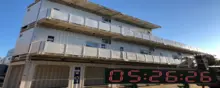 A multi-level structure made from stacked white shipping containers, featuring balconies and doors, set outdoors under a clear blue sky, with a digital timer display in the foreground.